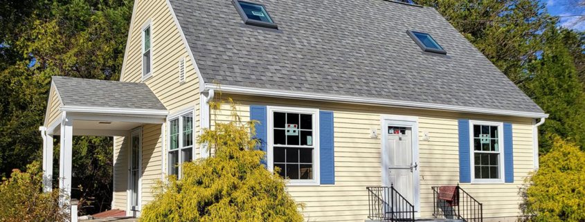 Exterior of a Cape Cod-style house featuring light yellow horizontal lap siding, a gray asphalt shingle roof with two skylights, and bright blue shutters. The front entrance has white stairs with a black metal railing and a white door, while a small white gabled porch is visible on the side of the house.