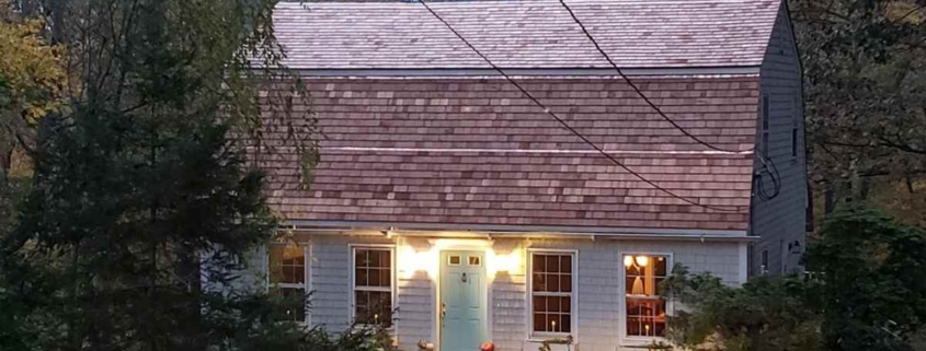 Exterior of a Cape Cod-style house featuring light gray cedar shingle siding and a steep gambrel roof with reddish-brown wood shingles. The front of the home is illuminated by warm interior lights, showcasing four white-trimmed windows and a bright light blue front door flanked by small decorative pumpkins.