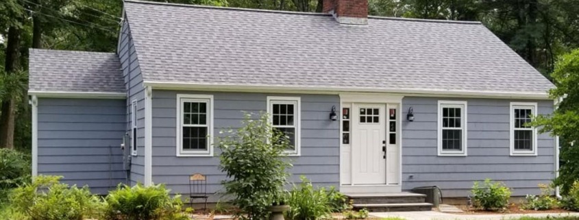 A single-story cottage with blue-gray horizontal lap siding, white-trimmed windows, a white front door with side lights, and a prominent red brick chimney.