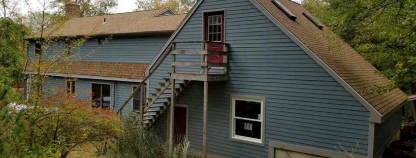 Exterior of a large, saltbox-style house featuring blue-gray horizontal lap siding and a brown asphalt shingle roof with skylights. The side of the home includes an external wooden staircase leading to a red door on the upper level, surrounded by dense trees and greenery.