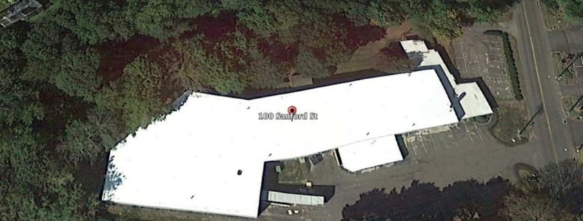 Aerial view of large building with flat white roof and parking.