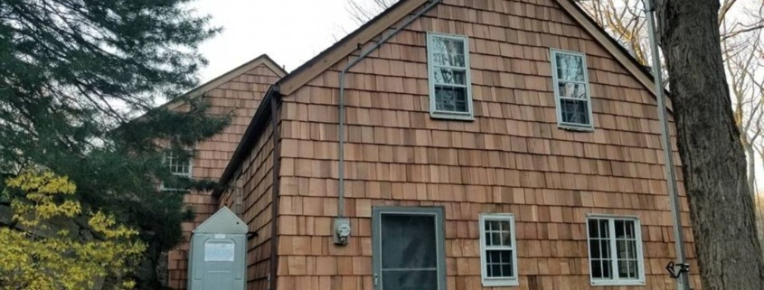 Exterior of a two-story house featuring natural wood cedar shingle siding, a steep gabled roof, and various white-trimmed windows, with a gray storm door serving as the primary entrance.