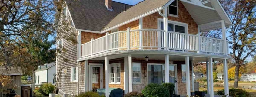 Two-story cedar shingle house with a large white balcony, gabled roof, and sunburst architectural detail.