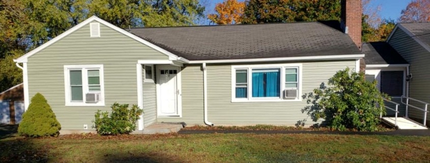 Single-story home with light sage green horizontal siding, white trim, a dark roof, and a brick chimney.