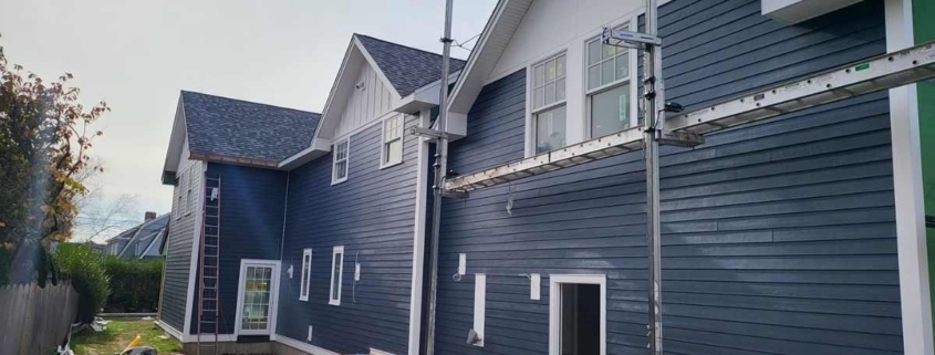 Exterior view of a home under construction featuring dark blue horizontal lap siding, white trim, and multiple white gables. Scaffolding and construction tools, including a miter saw and table saw, are visible in the yard along the side of the building.