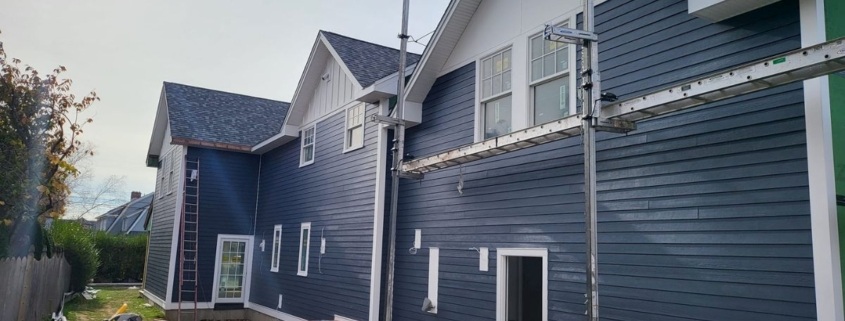 Exterior view of a home under construction featuring dark blue horizontal lap siding, white trim, and multiple white gables. Scaffolding and construction tools, including a miter saw and table saw, are visible in the yard along the side of the building.