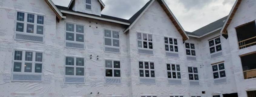 Exterior view of a multi-story building under construction with white weather-protective wrap, multiple rows of windows, and several prominent gables along the roofline.