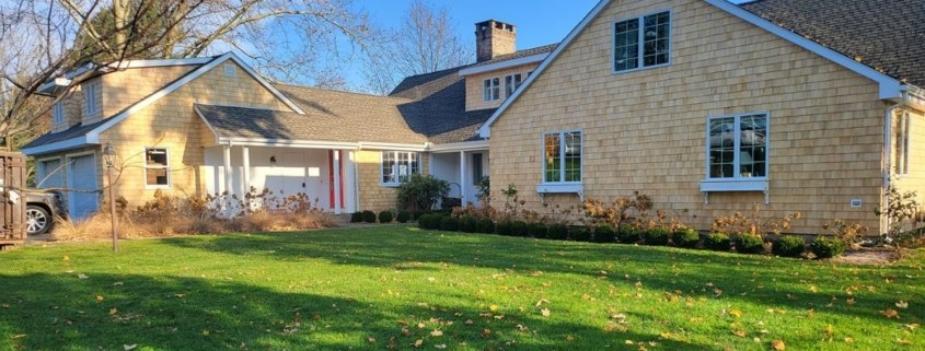 A large ranch-style home with light wood shingle siding, multiple gabled rooflines, white-trimmed windows with blue-gray planters, and a small red front door tucked under a covered walkway.