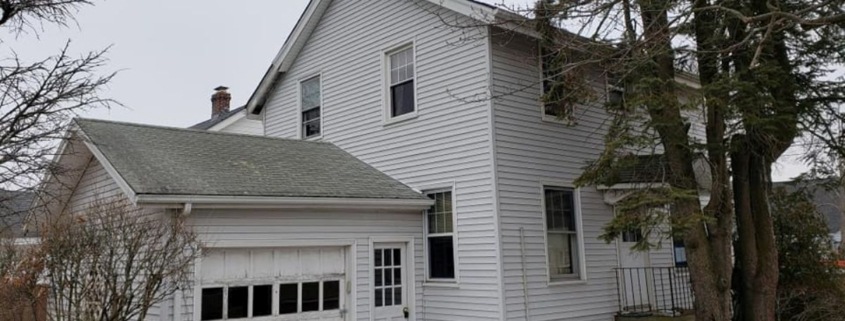 Exterior of a two-story house with white horizontal lap siding and a attached single-car garage, featuring patches of snow on the ground and large evergreen trees nearby.