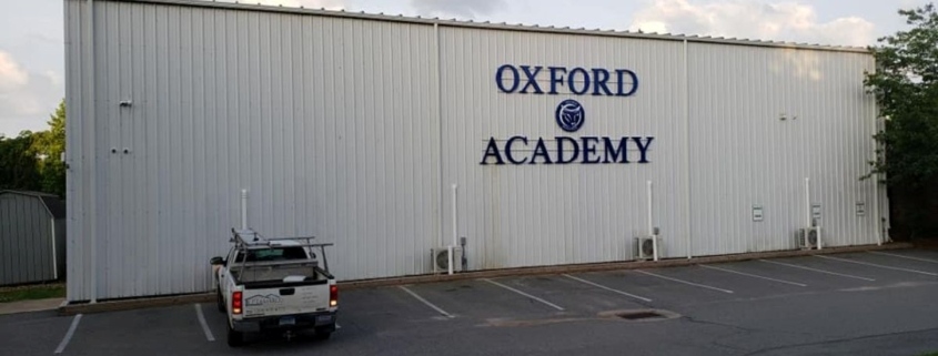 Exterior of a large commercial building with white vertical corrugated metal siding, featuring the blue "OXFORD ACADEMY" logo on the side. A white pickup truck with a ladder rack is parked in the foreground in a paved parking lot.