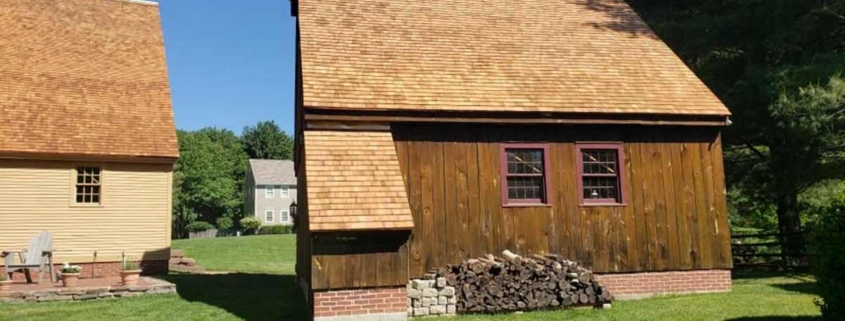 Exterior of a rustic outbuilding featuring vertical dark wood siding, a cedar shingle roof, and a red brick foundation, with a large stack of firewood piled against the side.