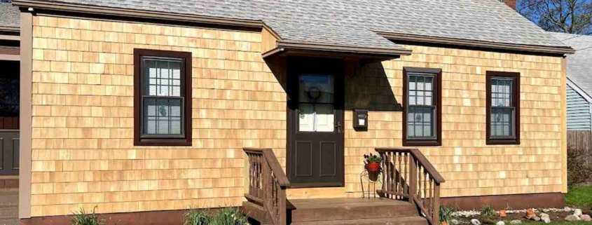 Small cottage-style house with light tan cedar shingle siding, dark brown trim around the windows and door, and a small wooden front porch with stairs.