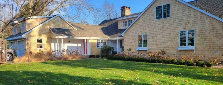 A sprawling ranch-style home featuring light yellow cedar shingle siding, white trim, and a prominent brick chimney, set behind a green lawn scattered with autumn leaves.
