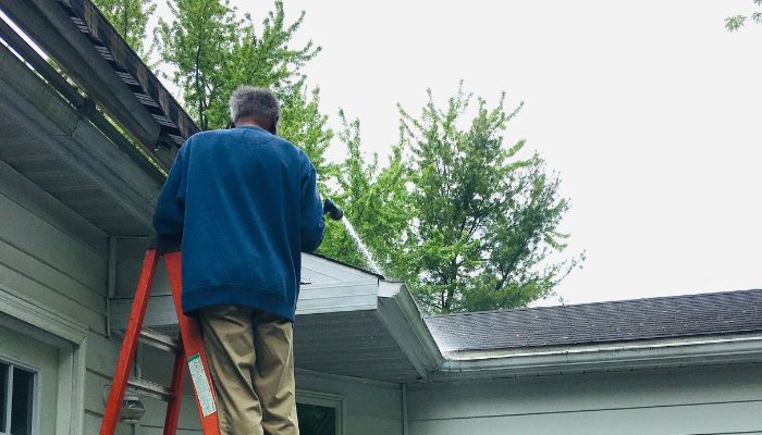 A person cleaning the roof gutters using a ladder and a hose on a residential home.