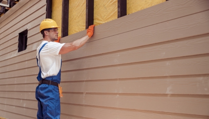 A man wearing overalls and a hard hat is painting house siding, representing labor costs for Hardie Plank siding in Connecticut.