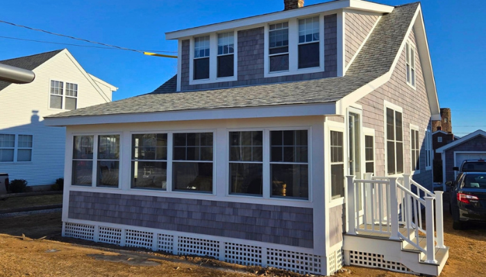 A finished coastal-style home featuring gray cedar shake siding, a large enclosed sunroom with white trim, and a matching lattice foundation.