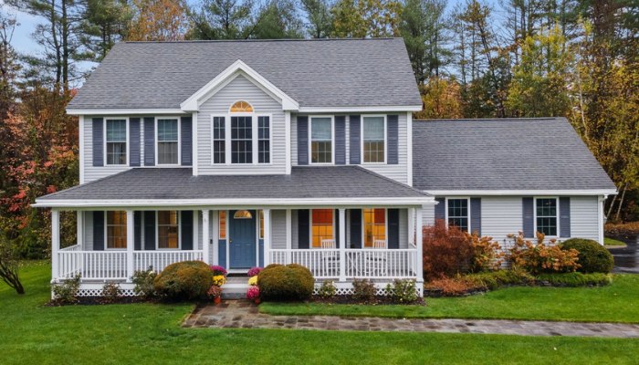 Two-story light gray house with a large wrap-around front porch, dark blue shutters, and a blue front door surrounded by autumn landscaping.