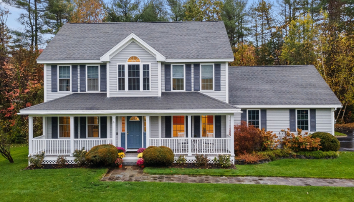 Two-story light gray house with a large wrap-around front porch, dark blue shutters, and a blue front door surrounded by autumn landscaping.