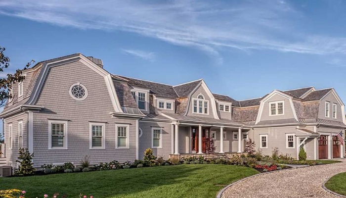 Large gray coastal-style estate with gambrel rooflines, cedar shake siding, and a white pillared porch along a gravel driveway.