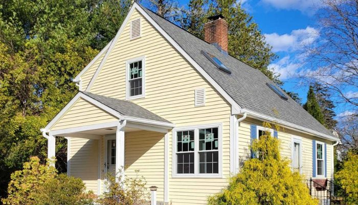 Small pale-yellow cottage with a steep gabled roof, white-trimmed windows, and blue shutters nestled among lush green trees.