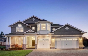 Two-story home with dark gray shingle gables, tan lap siding, white trim, and stone porch columns.
