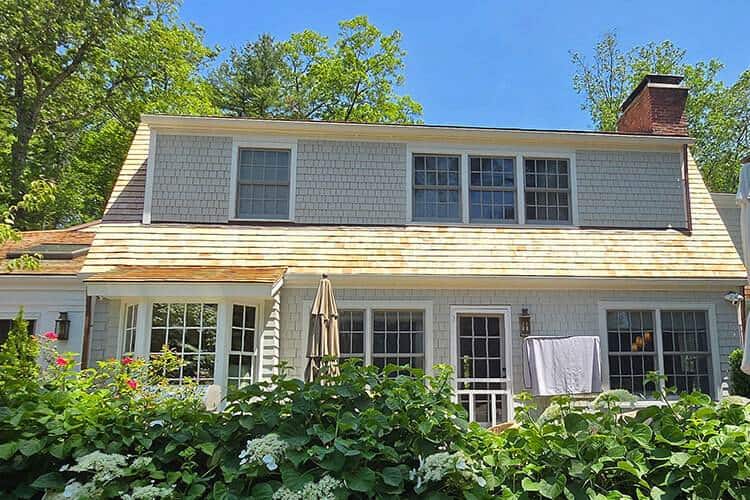 Two-story shingle house with a gambrel roof, dormer windows, and lush green bushes in front.
