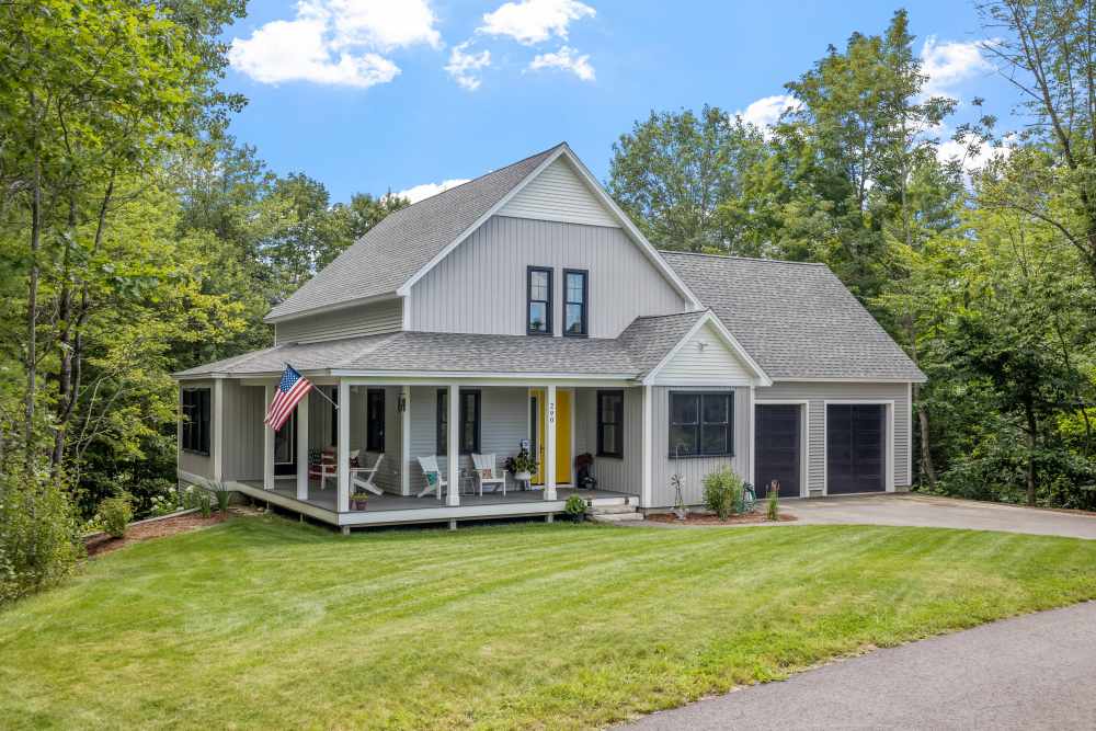 Light gray farmhouse with a wraparound porch, yellow front door, and a two-car garage.
